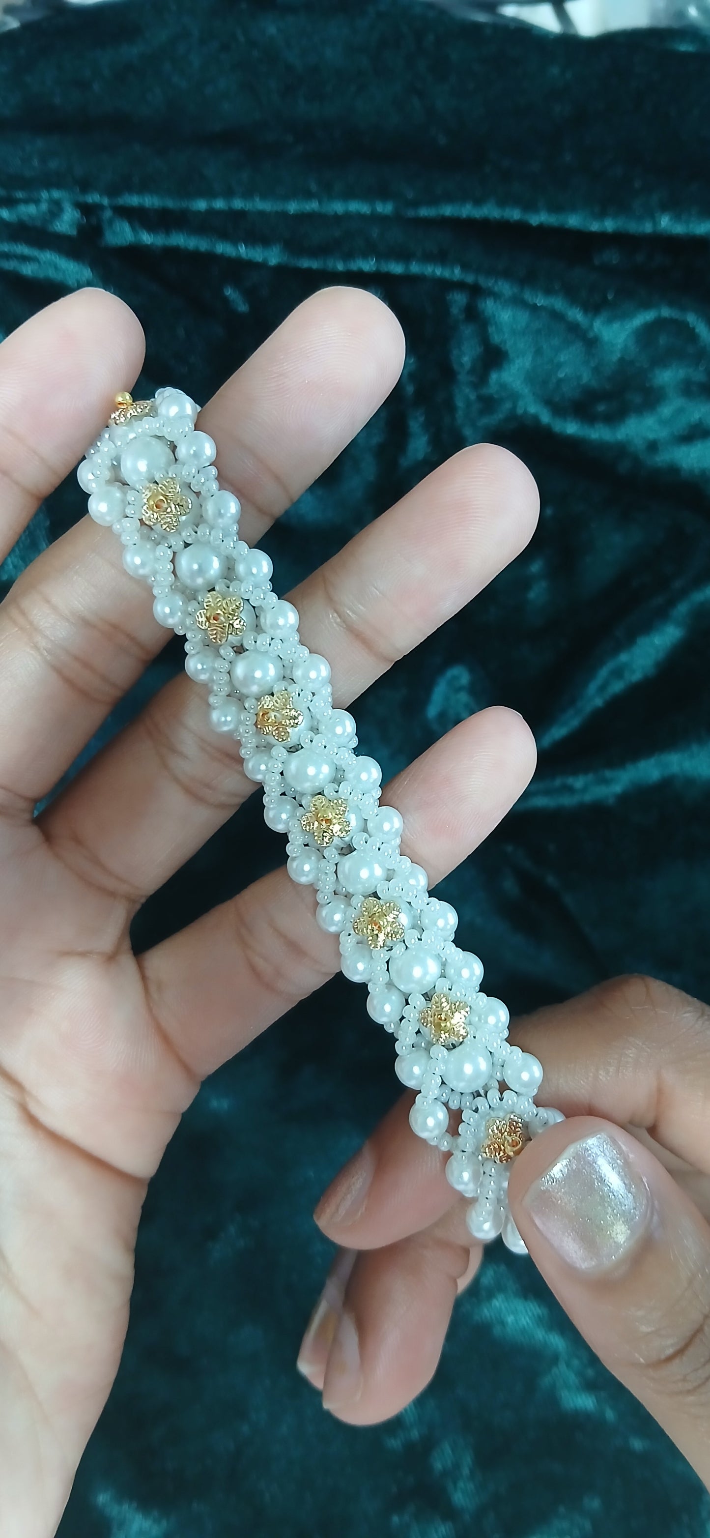 Hand holding a decorative bracelet with pearls and gold accents against a dark green velvet background.